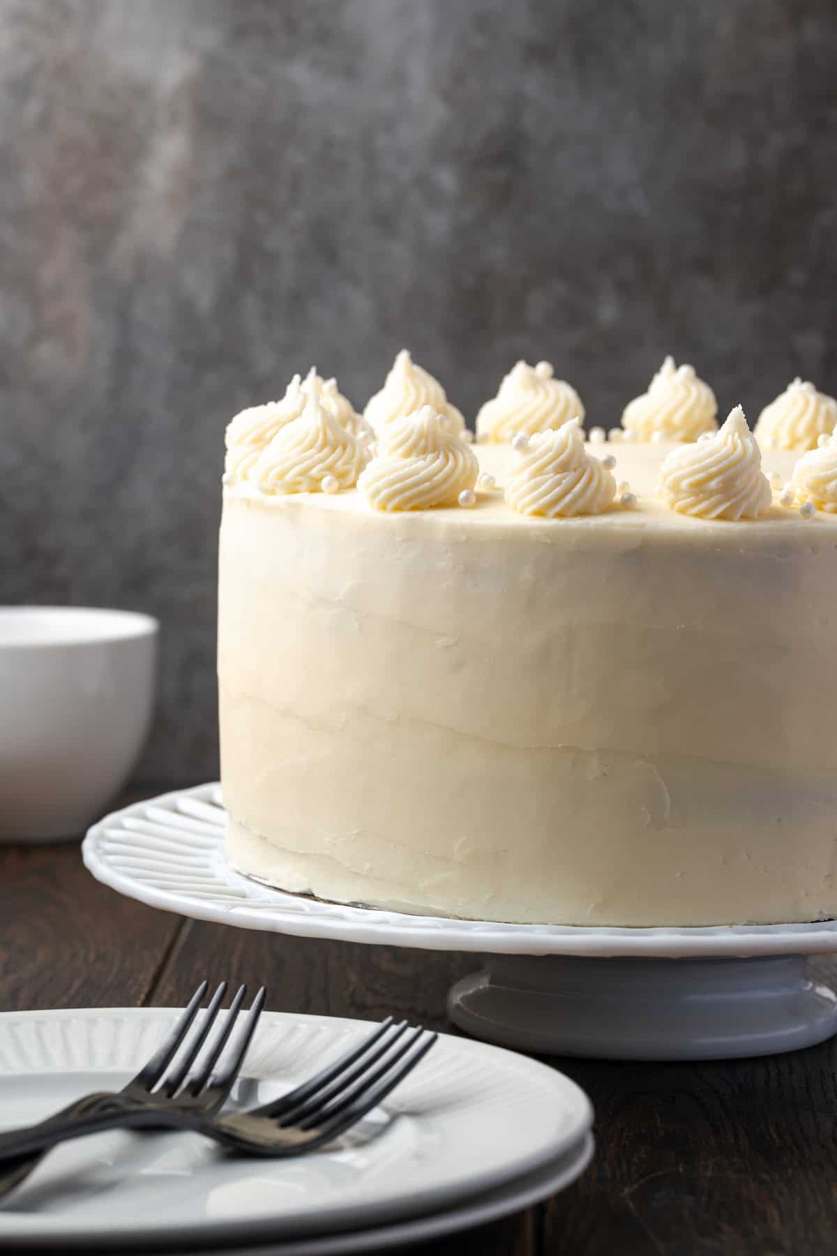 C'est le meilleur gâteau à la vanille à base de zéro! Apprenez à faire deux couches de gâteaux de vanille légères, moelleuses et parfaites givrées de crème au beurre. Vue latérale d'un gâteau de couche de vanille givrée sur un support à gâteau.