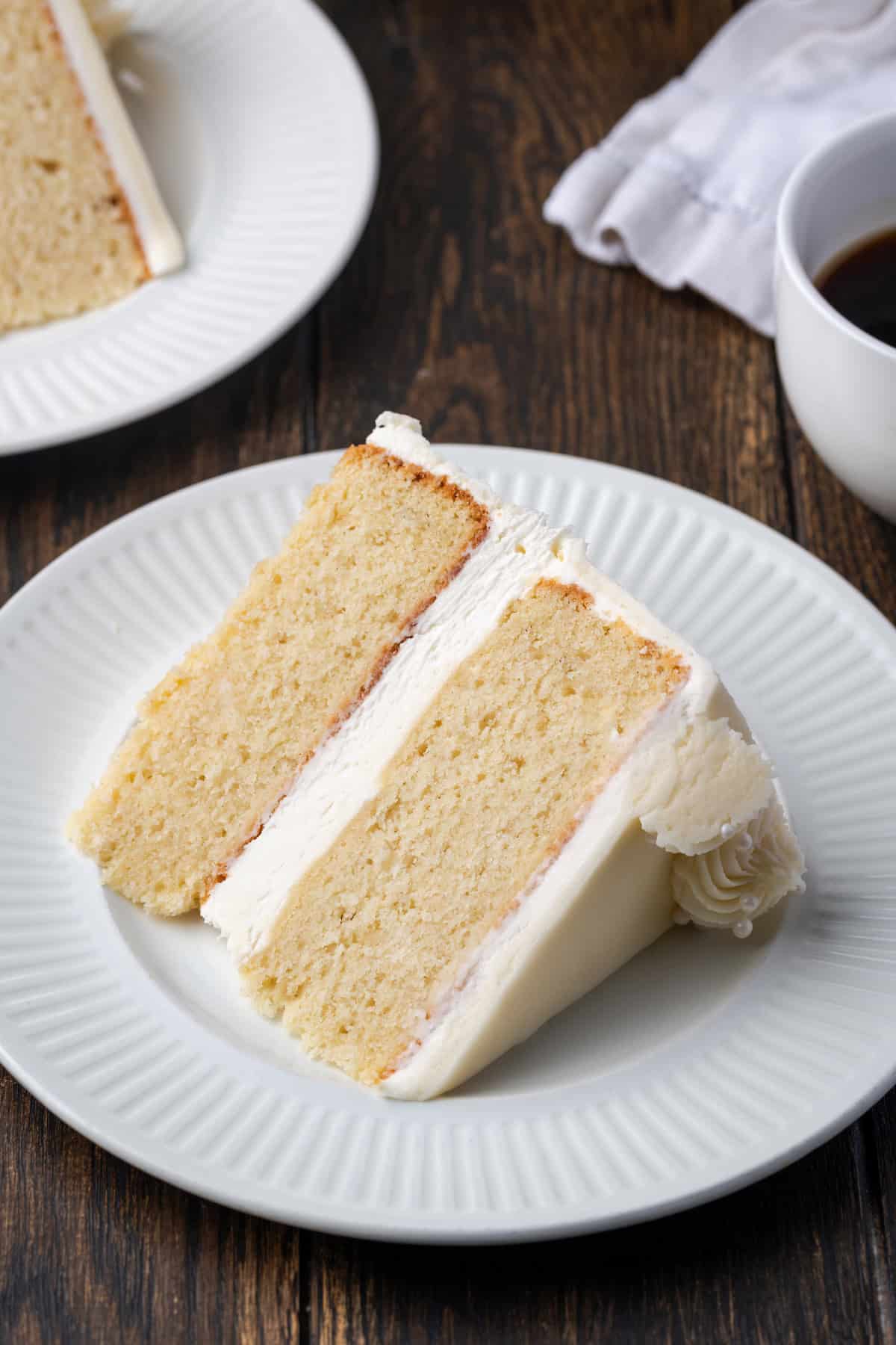 C'est le meilleur gâteau à la vanille à base de zéro! Apprenez à faire deux couches de gâteaux de vanille légères, moelleuses et parfaites givrées de crème au beurre. Une tranche de gâteau à la vanille sur une assiette blanche, avec une deuxième tranche en arrière-plan.