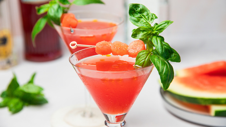 Deux verres à martini pastèque avec des bouteilles et des tranches en arrière-plan.